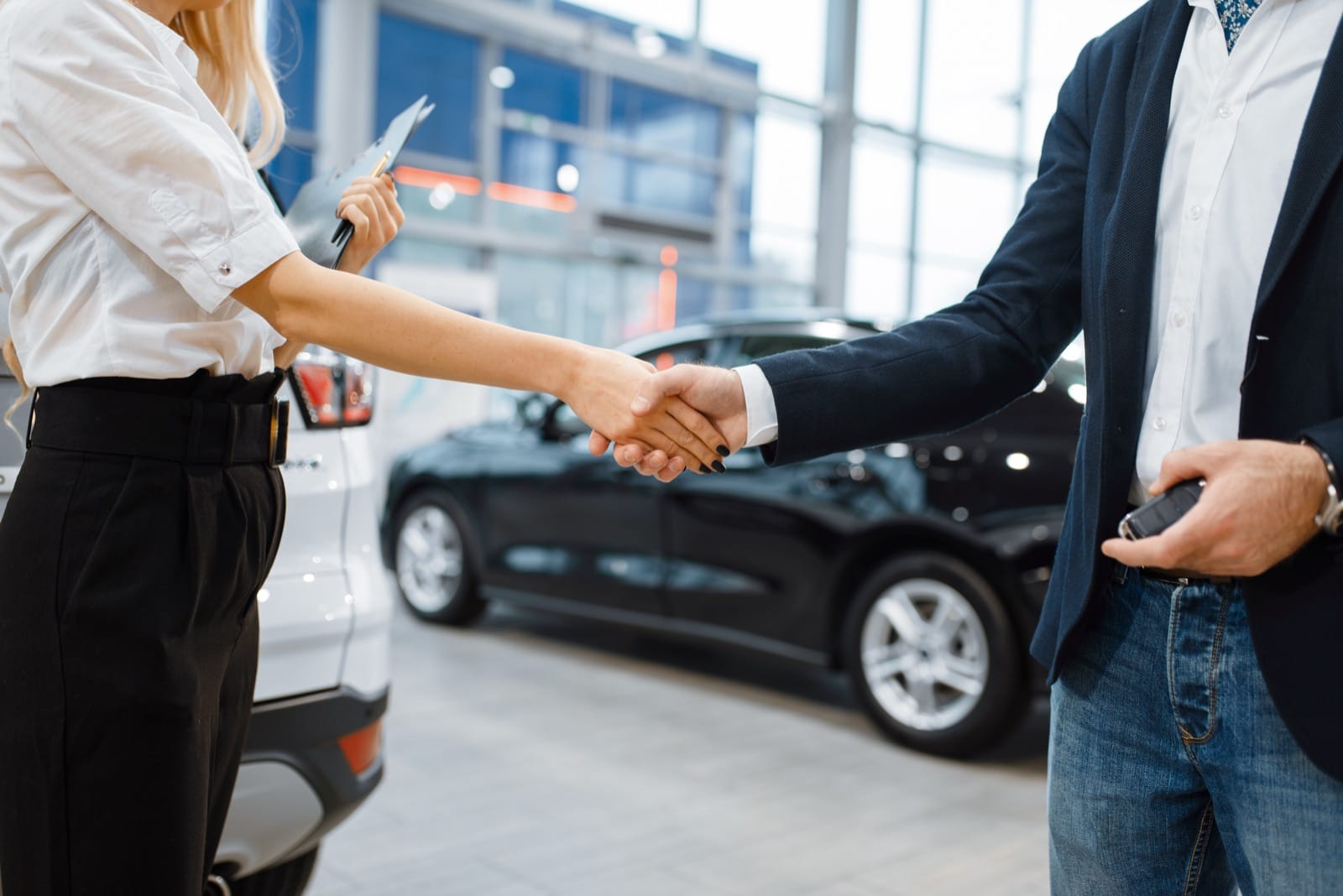 A broker and a client shaking hands in front of a car at a dealership